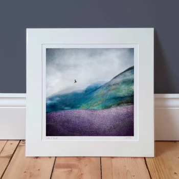a Hills of Tweed mounted print propped up against a dark grey wall. The print is an abstract picture of a Scottish hillside with a bird flying above in the distance. There is a tweed texture in the foreground.