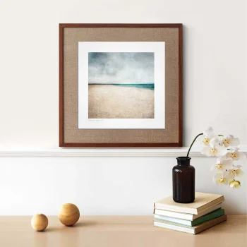 Kinshaldy Print in a brown frame on a cream wall. A vase on top of small books beside wooden ornaments shows an idea for home decor and how it can look in your house. Kinshaldy beach print is grey, greeny blue and sand coloured