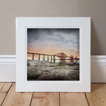 Queensferry Sunset mounted print against a beige wall. The picture is of the Forth Railway bridge with a sunset in the background and rocks in the foreground as it's from the South Queensferry side looking over to Fife.