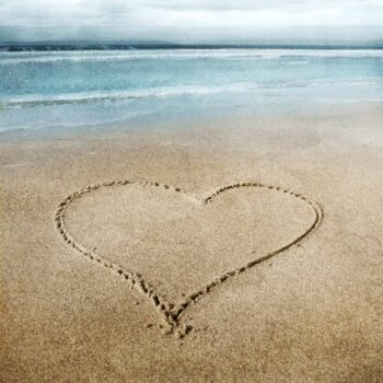 A heart carved in the sand, big in the foreground. In the background is ocean waves in blue/green.