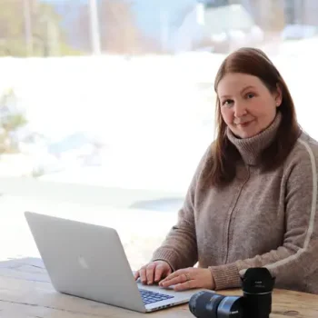 a photo of Carol McEwan, the artist, sitting at a chunky wooden table in front of a laptop, with camera lenses on the table. There's a snowy scene of trees and a house in front of a loch seen in the window behind.