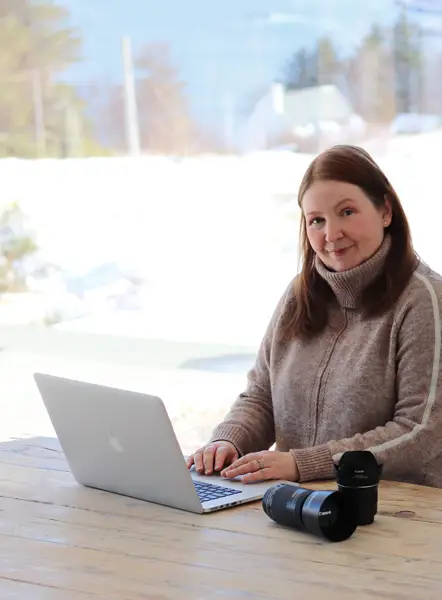 a photo of Carol McEwan, the artist, sitting at a chunky wooden table in front of a laptop, with camera lenses on the table. There's a snowy scene of trees and a house in front of a loch seen in the window behind.