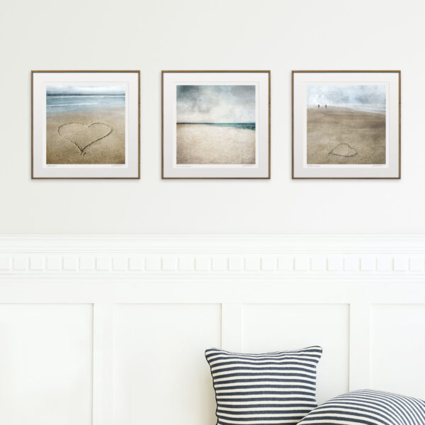 3 beach prints hanging on the wall in wooden frames in front of some cushions against a cream panelled wall