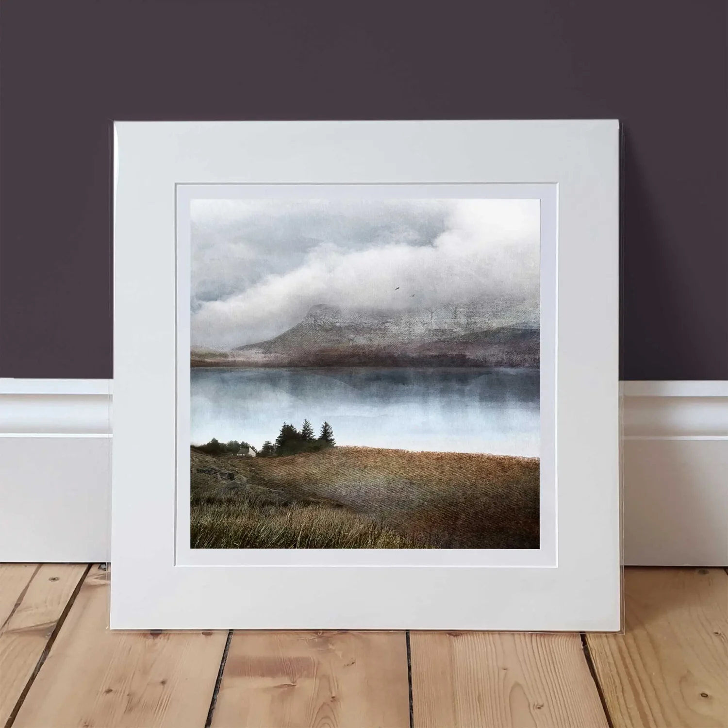 A Scottish, Highland view art print, of Little Loch Broom. There's a wee croft and trees in the foreground, with a silvery, blue loch and hen mountains in the background which fade into dramatic clouds. There's 2 birds in the sky just over the top of the mountains. The print is in a mounted window and is shown on a wooden floor with a background of a plum coloured wall.