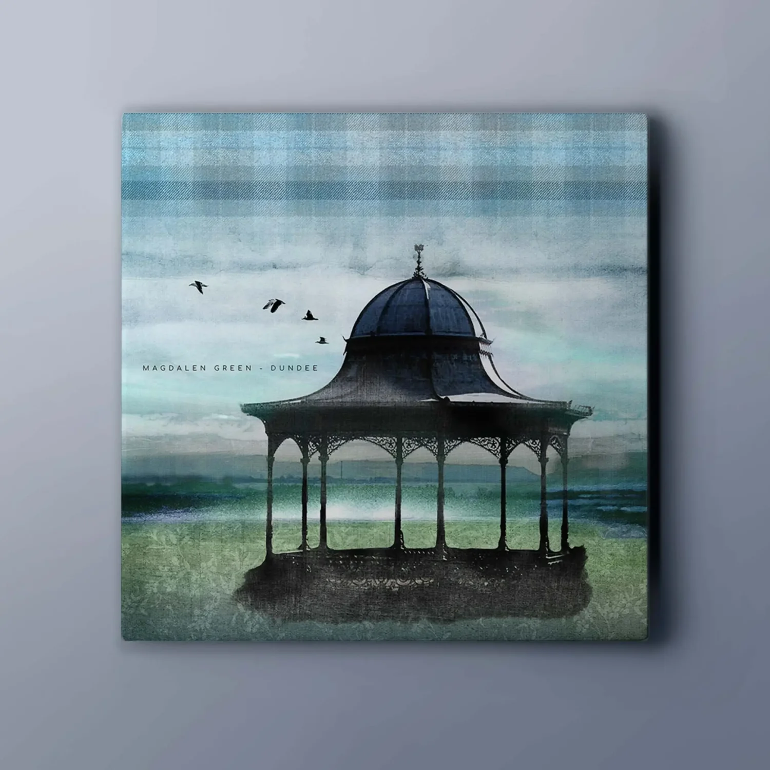 A grey wall with a canvas on it of a black bandstand (Magdalen GReen Dundee) and a tartan texture sky with birds inthe distance. There is text in the image that says Magdalen Green - dundee. The grass is green with a texture of white flowers and you can see the River Tay int eh background.