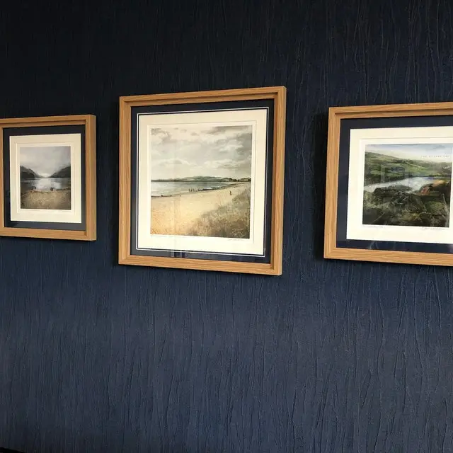 3 frames on a dark navy wall. The middle frame is larger than the other two and the frames are a light wood colour with a double mount