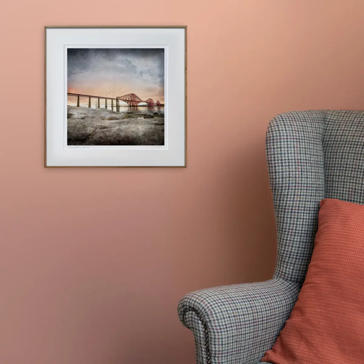 Queensferry Sunset print on a coral coloured wall. The picture is of the Forth Railway bridge with a sunset in the background and rocks in the foreground as it's from the South Queensferry side looking over to Fife. In the scene there is a tweed chair in front of the hanging framed print