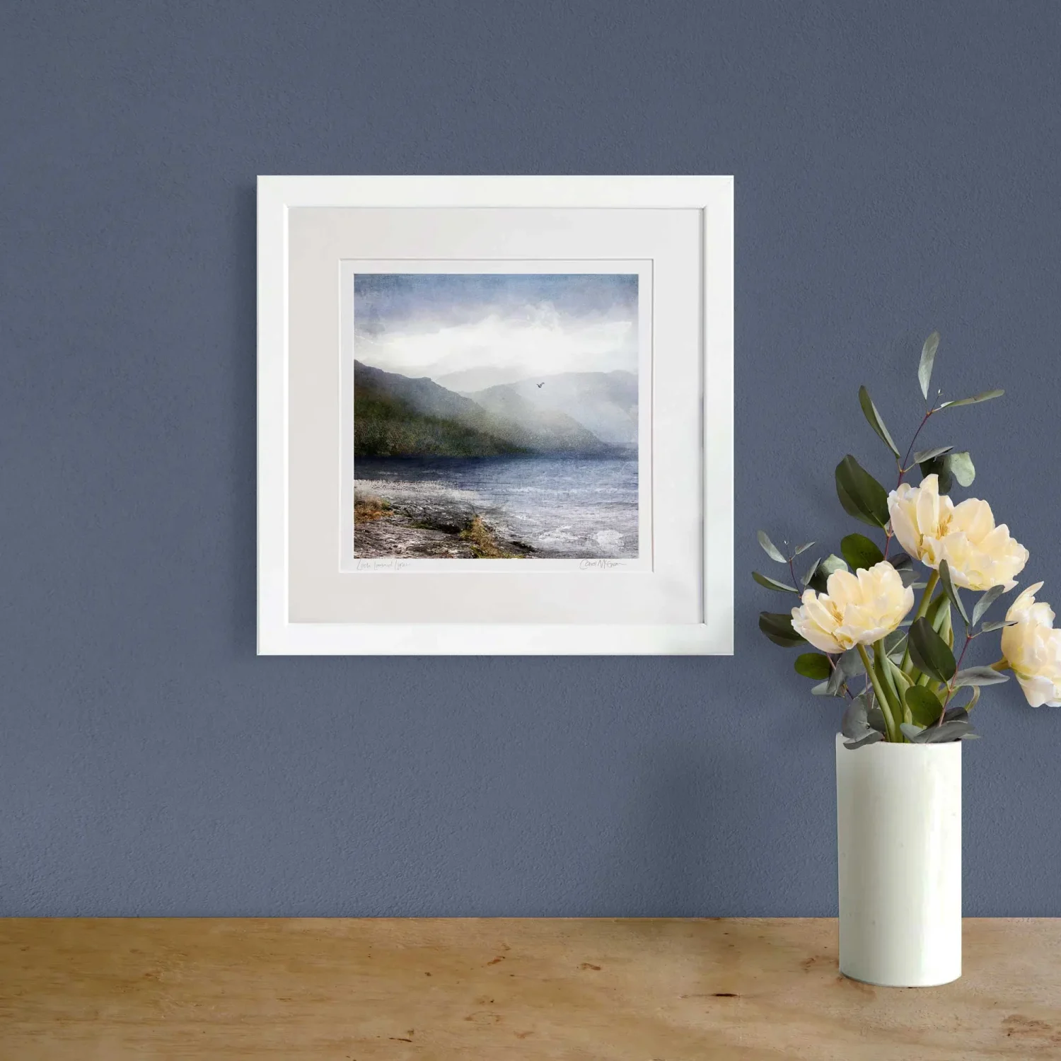 Loch Lomond art print in a white frame on a navy wall with a vase of flowers next to it on a wooden table