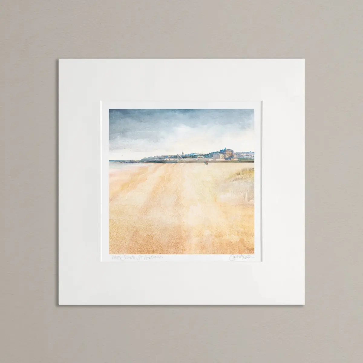 A mounted art print of West Sands, St Andrews, against a neutral grey background. The top 1/3 of the picture is the town shown as a view from the beach. The bottom part of the image is the beach. The sky is a muted blue grey. Overall the artwork is calming. There are 2 people walking in the distance on the beach